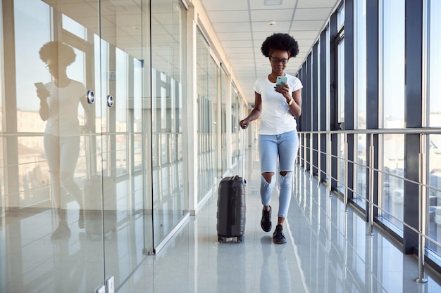 Junge afroamerikanische Passagierin in Freizeitkleidung ist mit Gepäck am Flughafen. Telefon verwenden.