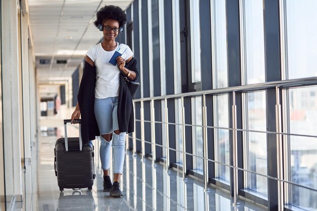 Junge afroamerikanische Passagierin in Freizeitkleidung und Kopfhörern ist mit Gepäck am Flughafen.