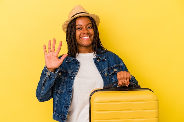 Junge afroamerikanische Reisende Frau mit Koffer auf gelbem Hintergrund isoliert lächelnd fröhlich zeigt Nummer fünf mit den Fingern.