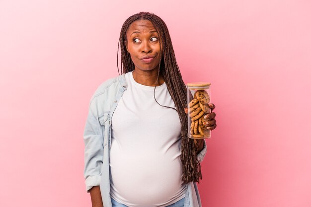 Junge afroamerikanische schwangere Frau, die Cookies einzeln auf rosafarbenem Hintergrund hält, verwirrt, fühlt sich zweifelhaft und unsicher.