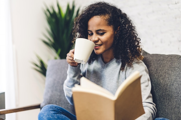 Junge afroamerikanische schwarze Frau, die trinkende Tasse heißen Kaffee oder Tee entspannt und Buch auf Couch zu Hause liest