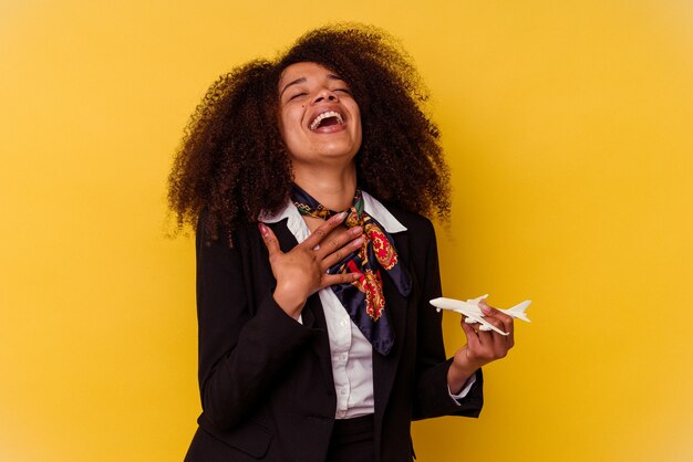 Junge afroamerikanische Stewardess, die ein kleines Flugzeug isoliert auf Gelb hält, lacht laut und hält die Hand auf der Brust.