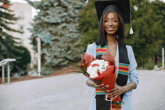 Junge afroamerikanische Studentin in blauer Kleidung und akademischer Mütze gekleidet. Mädchen posiert für ein Foto und hält Blumen