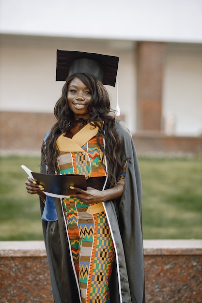 Junge afroamerikanische Studentin in schwarzem Abschlusskleid gekleidet. Campus als Hintergrund