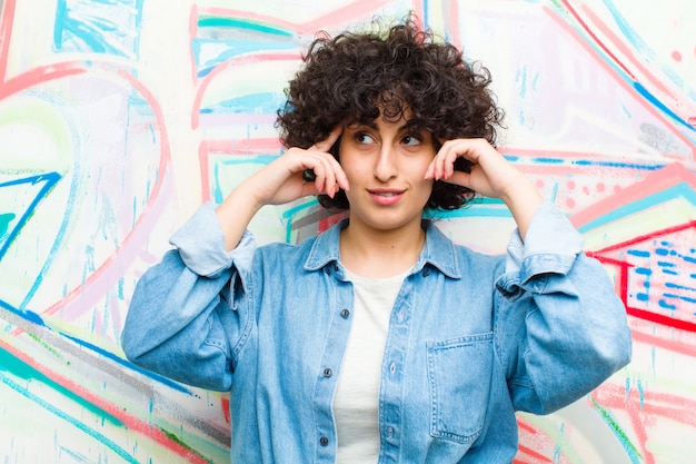 Junge Afrofrau, die verwirrt oder zweifelnd sich fühlt, auf eine Idee sich konzentriert, stark denkt und schaut, um Raum auf Seite auf Graffitiwand zu kopieren