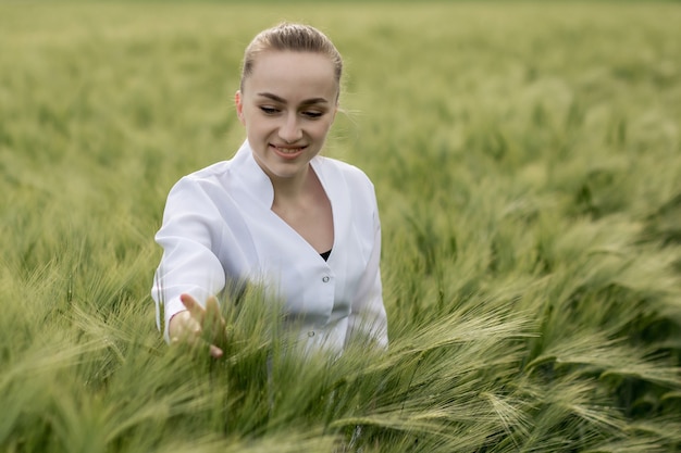 Junge Agronomenin im weißen Kittel, die im grünen Weizenfeld hocken und die Erntequalität überprüfen.