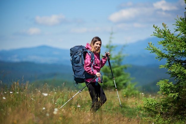 Junge aktive Kletterin mit Rucksack und Trekkingstöcken, Trekking in den Bergen, sonnigen Tag genießend. Konzept des aktiven Lebensstils