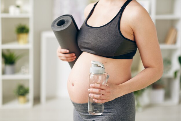 Junge aktive schwangere Frau mit gerollter Matte und Flasche mit reinem stillem Wasser stehend, während sie körperliche Übungen machen