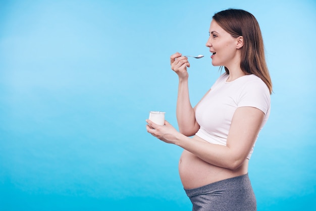 Junge aktive schwangere Frau mit kleinem Plastikbecher des Milchprodukts, das seinen Geschmack isoliert mit copyspace für Ihre Nachricht genießt