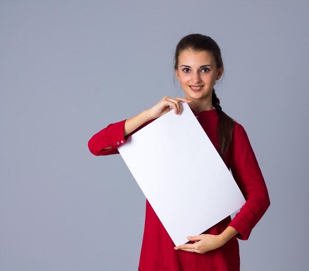 Junge angenehme Frau in Bluse mit Zopf mit weißem Blatt Papier auf grauem Hintergrund im Studio