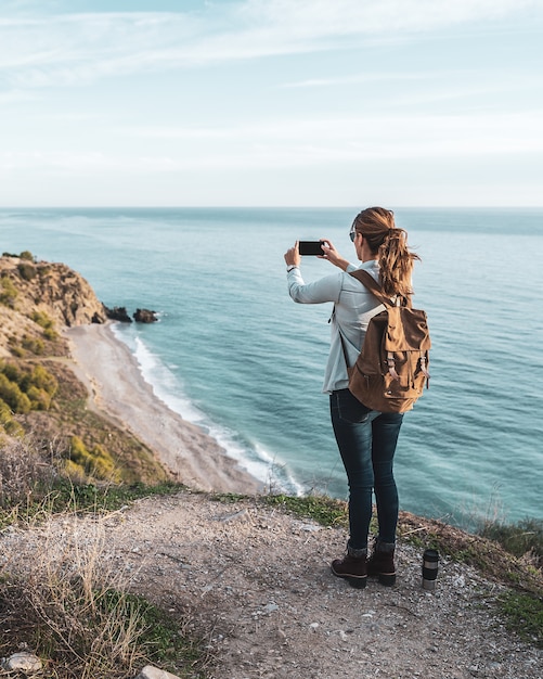 Junge angesagte Frau mit einem Rucksack die Küste an einem schönen Tag erforschend und fotografierend. Konzept der Erkundung und Abenteuer