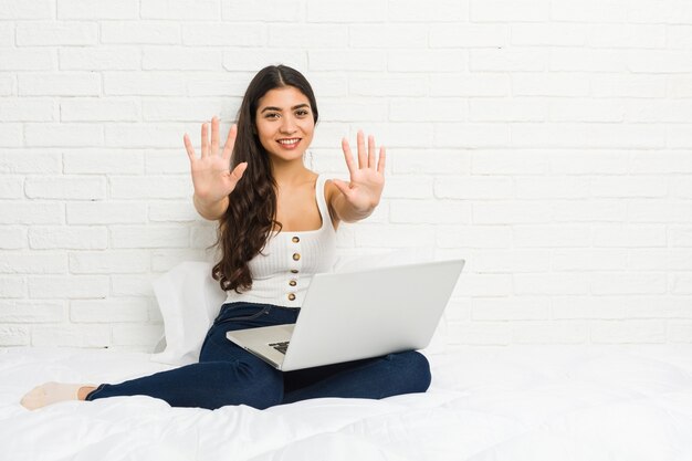 Junge arabische Frau, die mit ihrem Laptop auf dem Bett arbeitet, das Nummer zehn mit Händen zeigt.