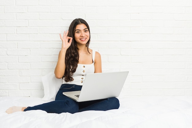 Junge arabische Frau, die mit ihrem Laptop auf dem Bett fröhlich und zuversichtlich arbeitet und ok Geste zeigt.