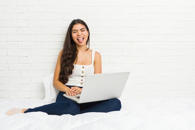 Junge arabische Frau, die mit ihrem Laptop auf dem Bett lustig und freundlich arbeitet, Zunge heraus haftend.