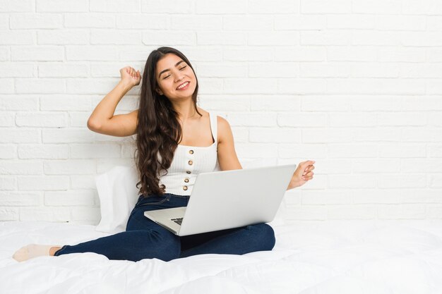Junge arabische Frau, die mit ihrem Laptop auf dem Betttanzen arbeitet und Spaß hat.