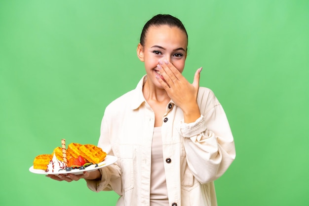 Junge arabische Frau, die Waffeln vor isoliertem Hintergrund hält, glücklich und lächelnd, den Mund mit der Hand bedeckend