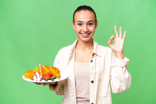 Junge arabische Frau, die Waffeln vor isoliertem Hintergrund hält und mit den Fingern ein Ok-Zeichen zeigt