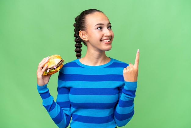 Junge arabische Frau hält einen Burger vor isoliertem Hintergrund und zeigt auf eine tolle Idee