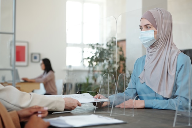 Junge arabische Frau in Hijab und Maske, die am Tisch mit Barriereschirm sitzt und die Staatsbürgerschaft beantragt