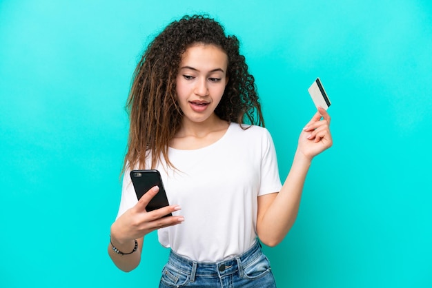 Junge arabische Frau isoliert auf blauem Hintergrund, die mit dem Handy mit einer Kreditkarte kauft