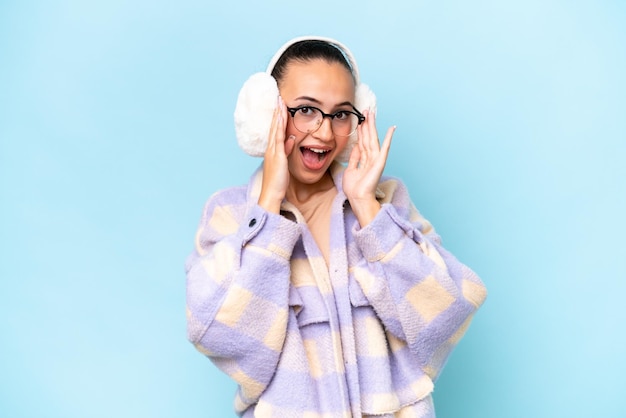 Junge arabische Frau mit Wintermuffs isoliert auf blauem Hintergrund mit Brille und überrascht