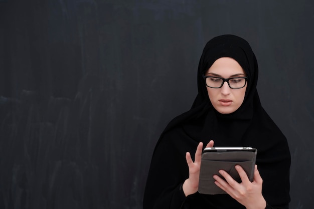 Junge arabische Geschäftsfrau in traditioneller Kleidung oder Abaya und Brille mit Tablet-Computer vor schwarzer Tafel, die moderne islamische Mode und Technologie darstellt