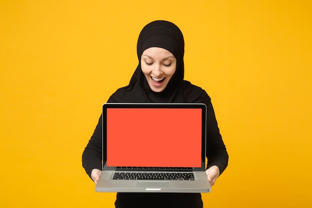 Junge arabische muslimische Angestellte Frau in Hijab schwarzer Kleidung halten und arbeiten Laptop-PC isoliert auf gelbem Wandporträt. Menschen religiöses Lifestyle-Konzept.