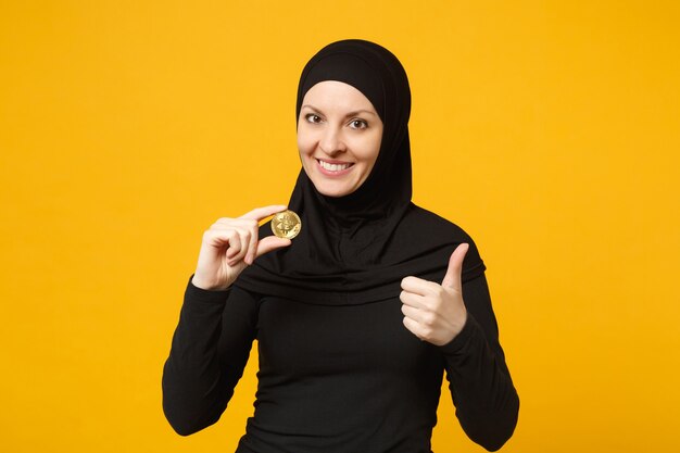 Junge arabische muslimische frau in hijab-schwarzer kleidung halten in der hand, die bitcoin-Münzenwährung isoliert auf gelbem wandporträt hält. Menschen religiöses Lifestyle-Konzept.