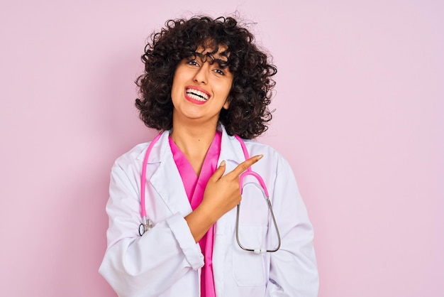 Junge arabische Ärztin mit lockigem Haar und Stethoskop vor isoliertem rosa Hintergrund, fröhlich