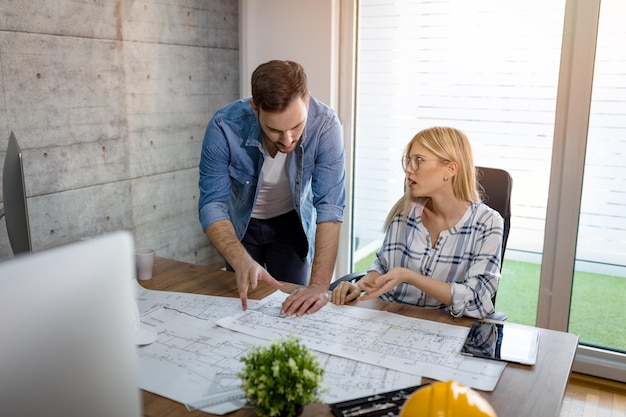 Junge Architektenkollegen unterhalten sich und analysieren Pläne, während sie im Büro an einem neuen Projekt arbeiten.