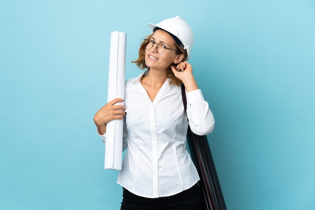 Junge Architektin Georgian Frau mit Helm und hält Blaupausen über isoliertem Hintergrund, der Zweifel hat