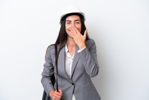 Junge Architektin, kaukasische Frau mit Helm und Blaupausen isoliert auf weißem Hintergrund, glücklich und lächelnd, den Mund mit der Hand bedeckend