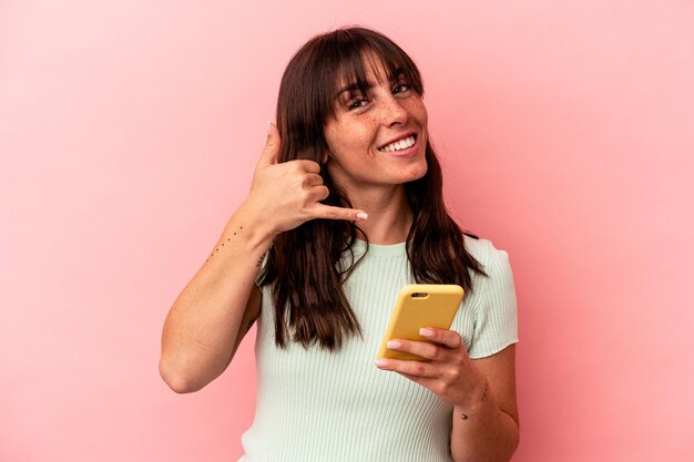 Junge argentinische Frau, die ein Mobiltelefon einzeln auf rosafarbenem Hintergrund hält, das eine Handyanrufgeste mit den Fingern zeigt.