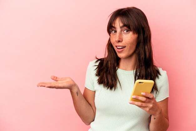 Junge argentinische Frau, die ein Mobiltelefon einzeln auf rosafarbenem Hintergrund hält, das einen Kopienraum auf einer Handfläche zeigt und eine andere Hand an der Taille hält.