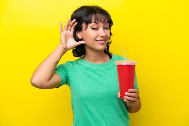 Junge argentinische Frau, die ein Soda isoliert auf gelbem Hintergrund hält und etwas hört, indem sie die Hand auf das Ohr legt