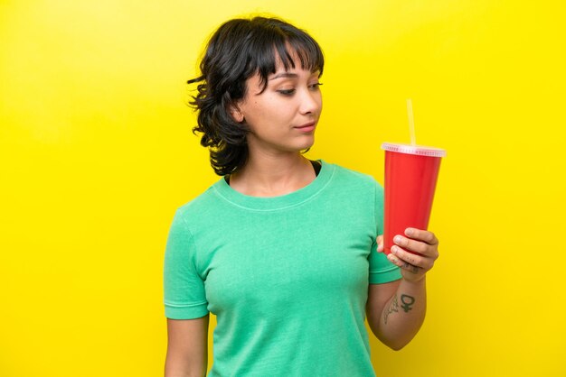 Junge argentinische Frau, die ein Soda isoliert auf gelbem Hintergrund hält und zur Seite schaut