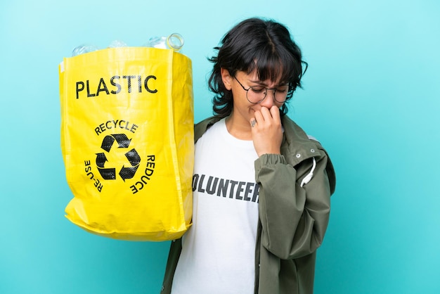 Junge argentinische Frau, die eine Tasche voller Plastikflaschen hält, um isoliert auf blauem Hintergrund zu recyceln, die Zweifel hat