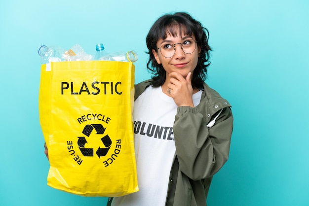 Junge argentinische frau, die eine tasche voller plastikflaschen hält, um isoliert auf blauem hintergrund zu recyceln, die zweifel hat