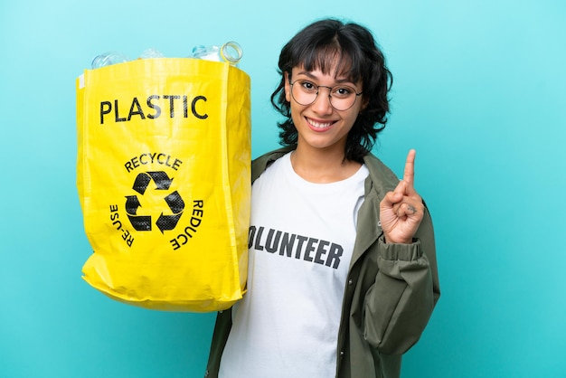 Junge argentinische Frau, die eine Tüte voller Plastikflaschen hält, um isoliert auf blauem Hintergrund zu recyceln, die einen Finger zeigt und hebt