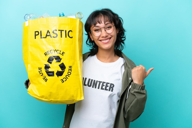 Junge argentinische Frau, die eine Tüte voller Plastikflaschen hält, um isoliert auf blauem Hintergrund zu recyceln, die zur Seite zeigt, um ein Produkt zu präsentieren