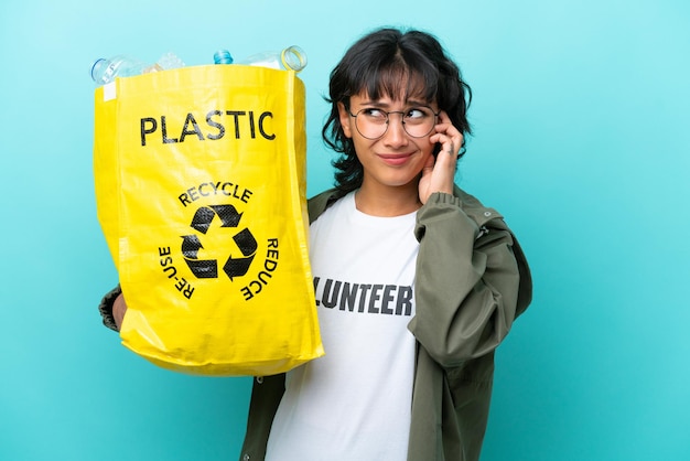 Junge argentinische Frau, die eine Tüte voller Plastikflaschen hält, um sie isoliert auf blauem Hintergrund zu recyceln, frustriert und bedeckt die Ohren