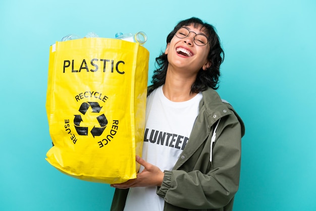 Junge argentinische Frau, die eine Tüte voller Plastikflaschen hält, um sie isoliert auf blauem Hintergrund zu recyceln und zu lachen