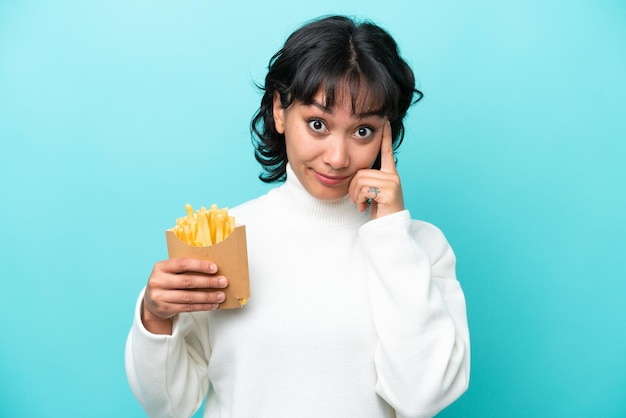 Junge argentinische Frau, die gebratene Chips isoliert auf blauem Hintergrund hält und eine Idee denkt