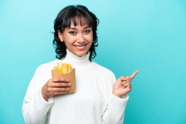 Junge argentinische Frau, die gebratene Chips isoliert auf blauem Hintergrund hält und mit dem Finger zur Seite zeigt