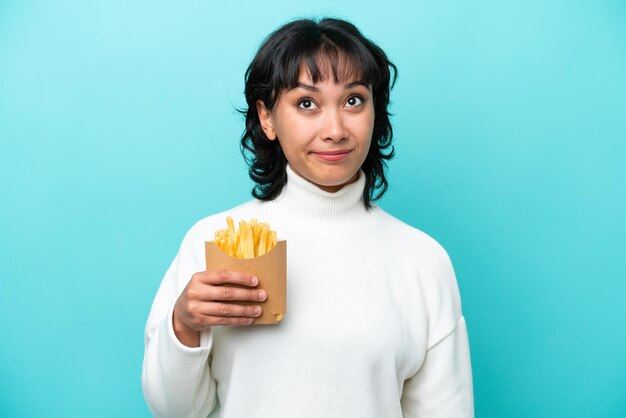 Junge argentinische Frau, die gebratene Chips isoliert auf blauem Hintergrund hält und nach oben schaut