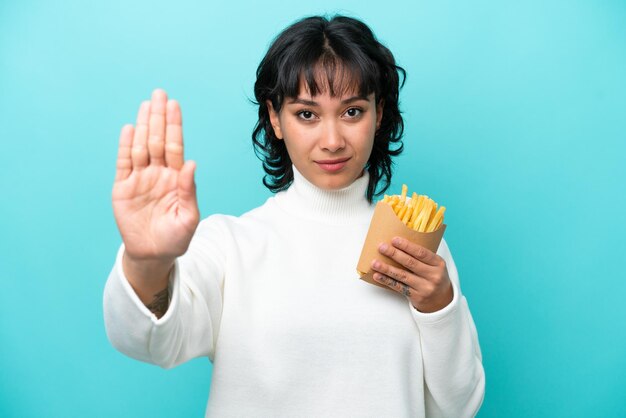 Junge argentinische Frau, die gebratene Chips isoliert auf blauem Hintergrund hält und Stoppgeste macht