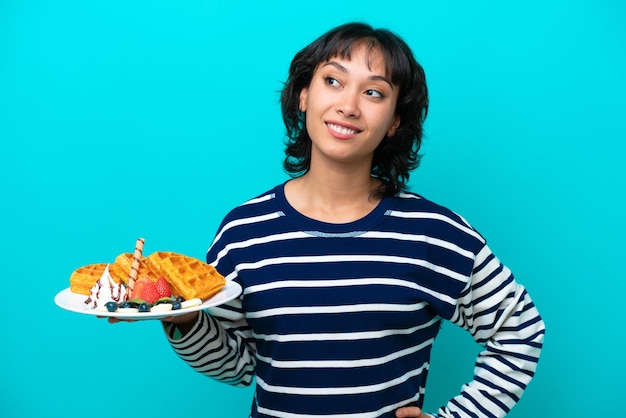 Junge argentinische Frau, die Waffeln isoliert auf blauem Hintergrund hält und eine Idee denkt, während sie nach oben schaut