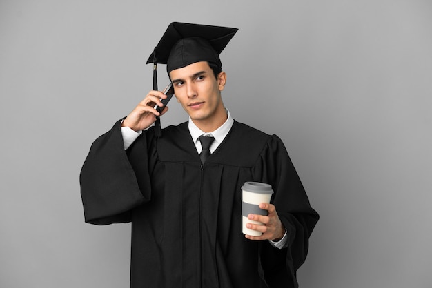 Junge argentinische Hochschulabsolventin isoliert auf grauem Hintergrund mit Kaffee zum Mitnehmen und einem Handy