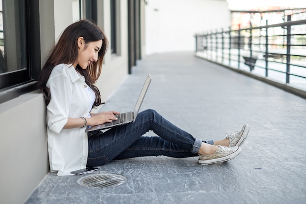 Junge Asiatin, die auf der Straße sitzt und mit ihrem Laptop bei der Unterhaltung am Handy arbeitet