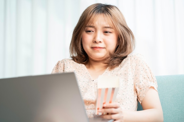 Junge Asiatin sitzt auf dem Sofa und isst und schaut Filme auf dem Laptop